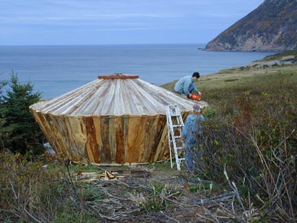 A man works on the roof of a wooden yurt in a hillside field overlooking the ocean.